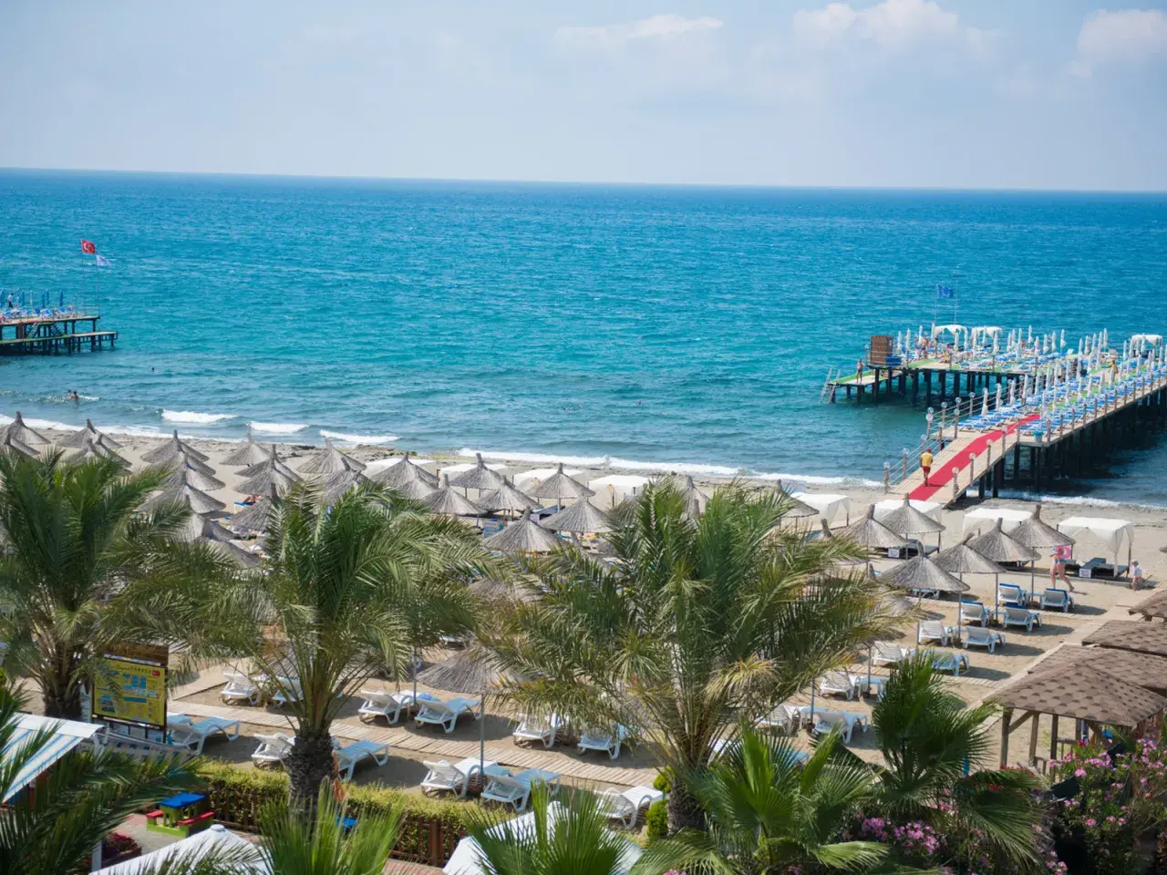 Sonne, Strand und Meer am quattro beach spa & resort. Liegestühle unter Strohschirmen, Palmen und ein langer Steg mit rotem Teppich laden zum Entspannen ein.