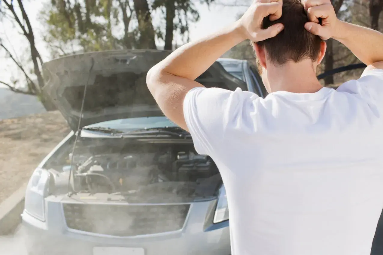 Hombre frustrado mira cómo sale humo blanco del motor de su coche averiado.