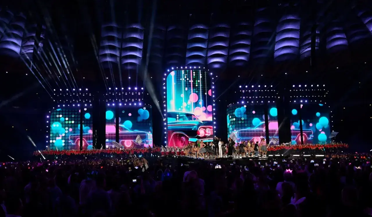 Wydarzenia na Stadionie Narodowym: scena z neonowymi samochodami i tańczącymi artystami, oświetlona przez reflektory. Tłum widzów wpatrzony w widowisko.