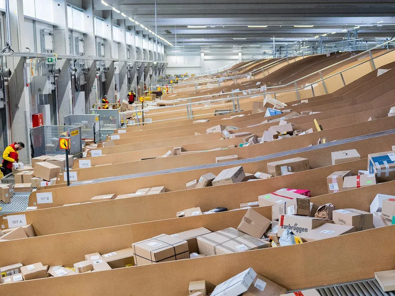 Arbeiter sortieren Pakete in einem riesigen DHL-Zentrallager. Zahlreiche Pakete liegen auf Förderbändern und in Regalen.