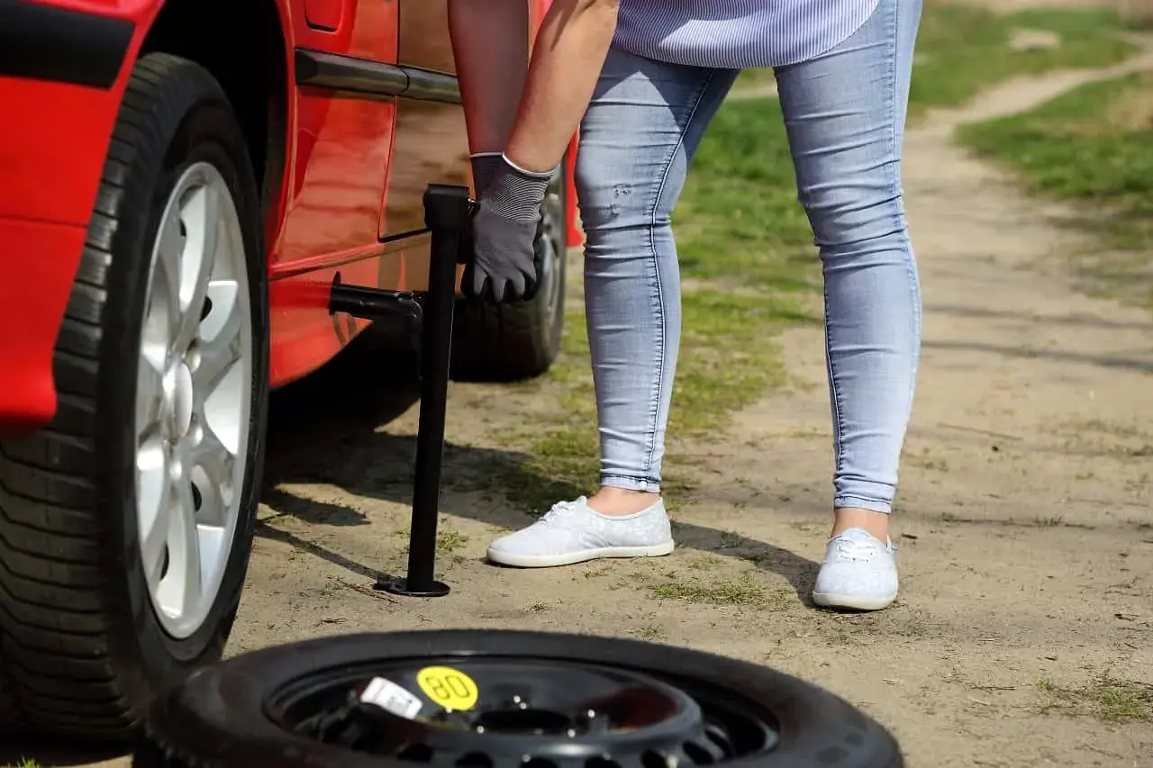 Kobieta wymienia koło w czerwonym samochodzie. Obok leży opona zapasowa z żółtą naklejką "80". Zastanawiasz się, ile można jeździć na oponie zapasowej?