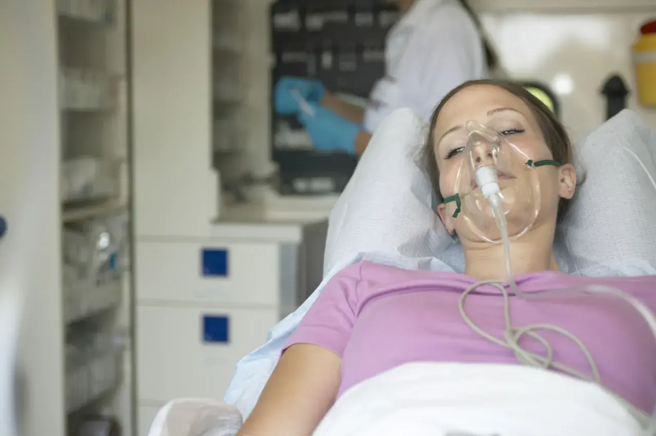 Młoda kobieta w masce tlenowej, leżąca w łóżku szpitalnym. Jej stan sugeruje chorobę popromienną, wymagającą intensywnej opieki medycznej.