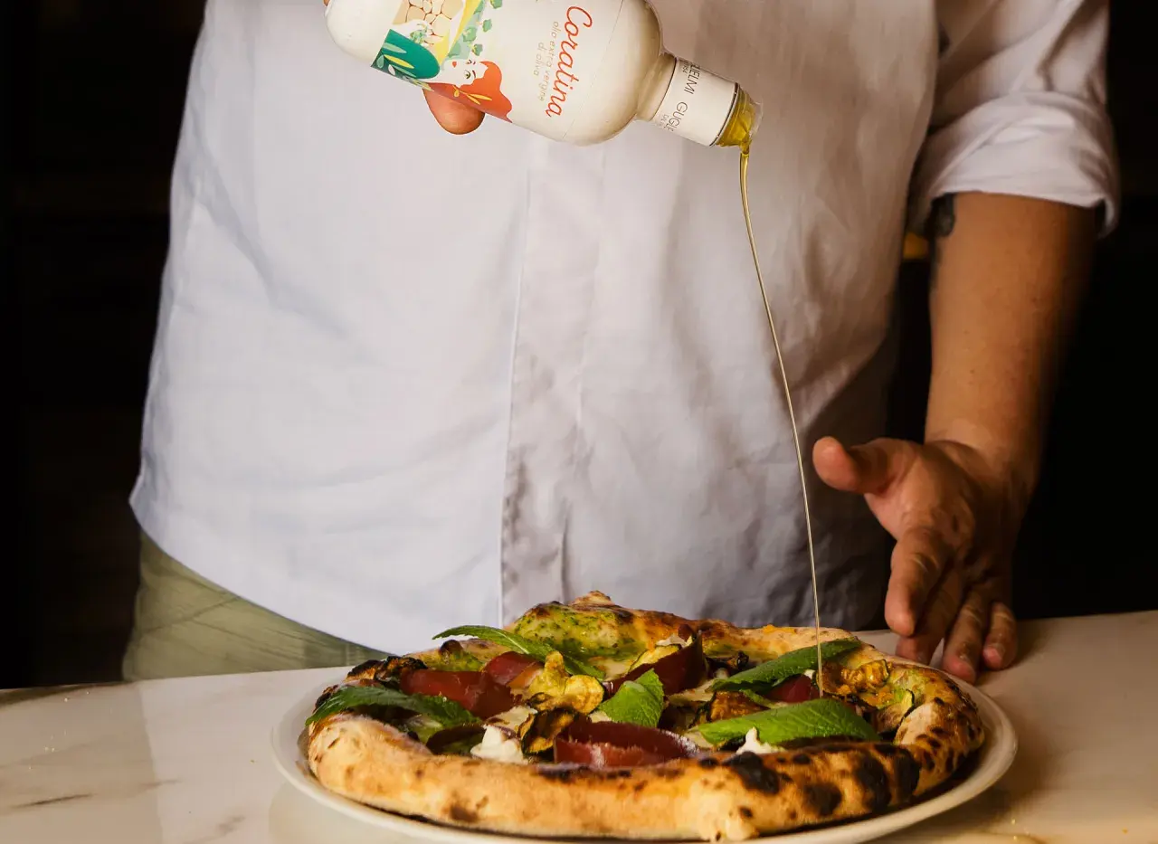 Un cuoco versa l'olio Coratina migliore su una pizza gourmet. L'olio extra vergine d'oliva esalta i sapori.