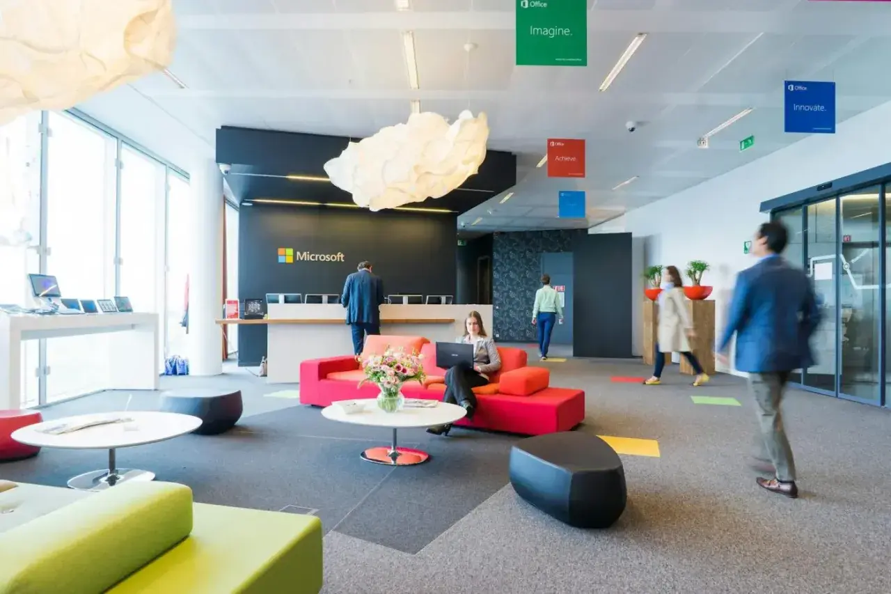 Un **open space** moderne chez Microsoft. Une femme travaille sur son ordinateur portable, entourée de canapés colorés. Des affiches "Imagine", "Achieve", "Innovate" flottent au plafond.