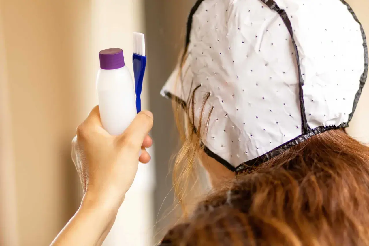 Persona se prepara para hacerse mechas en casa. Sostiene un kit para teñir el cabello y lleva un gorro especial.