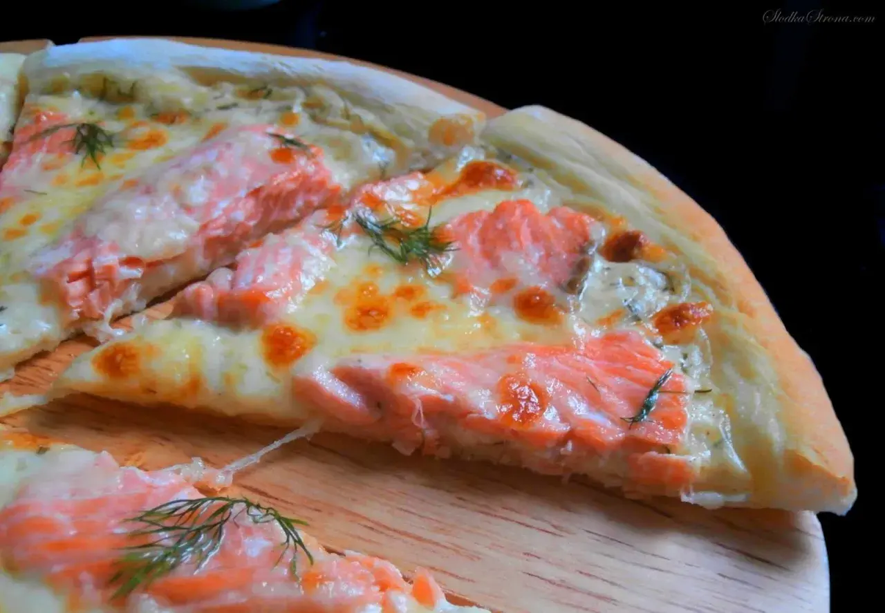Kawałek pysznej pizzy z łososiem, roztopionym serem i koperkiem na drewnianej desce.