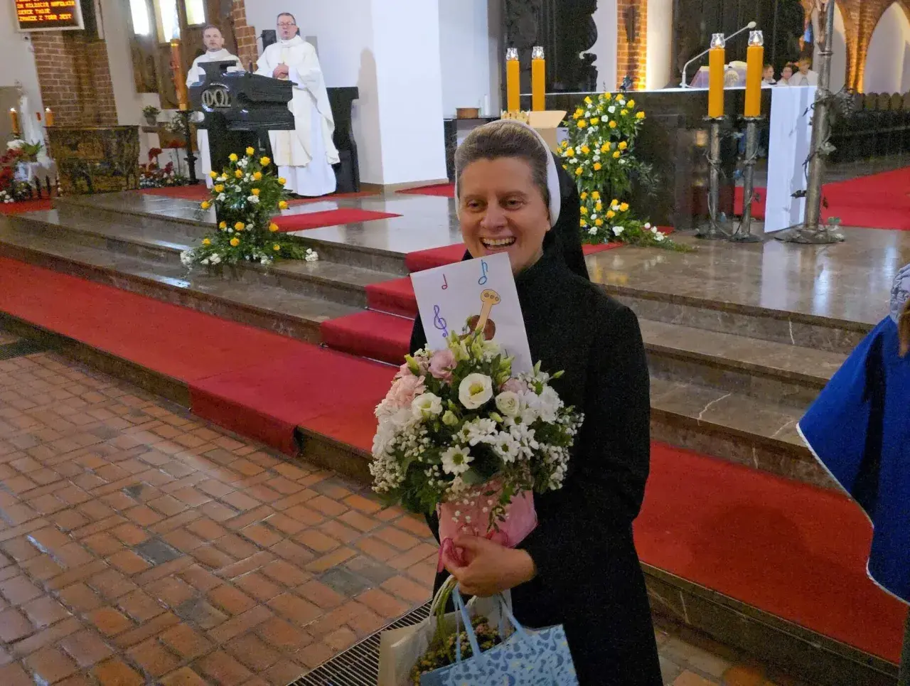 Radosna zakonnica z bukietem kwiatów i kartką z nutami. W tle msza święta, 25 października.