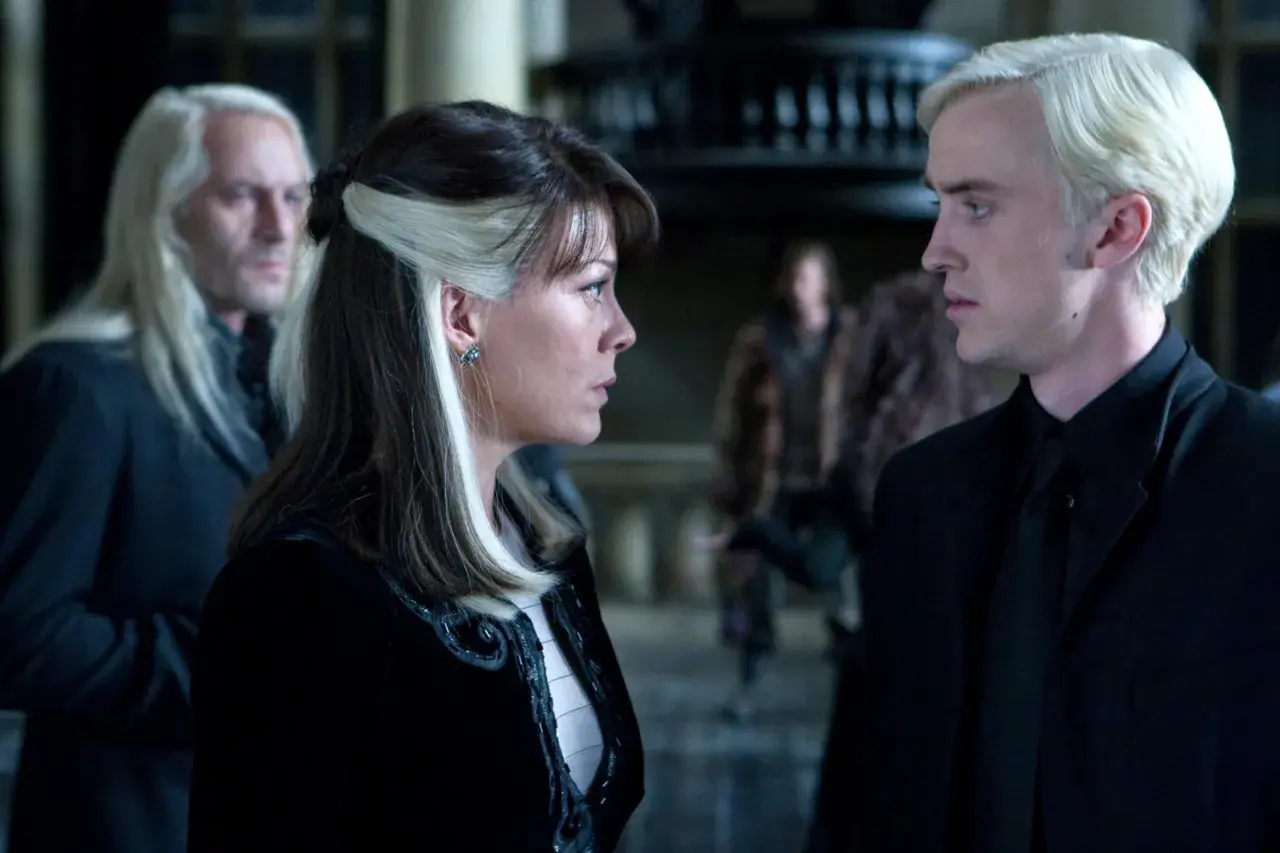 Narcyza Malfoy rozmawia z synem, Draco, w ponurym wnętrzu. W tle widać Lucjusza Malfoya.