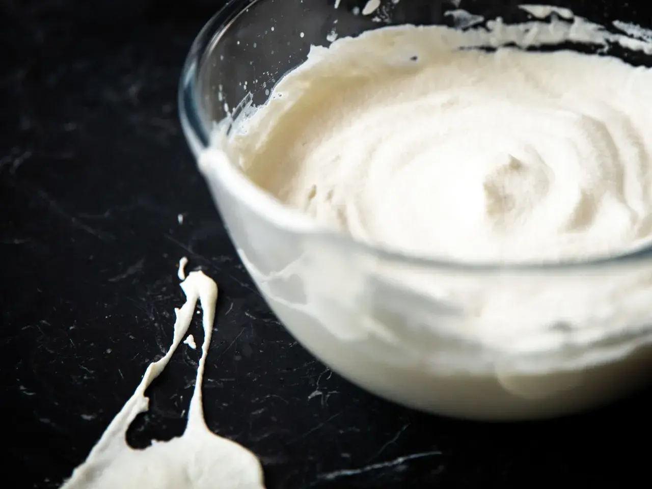 Schlagrahm in einer Glasschüssel, bereit für ein Dessert. Ein Klecks Sahne ist daneben auf der dunklen Arbeitsfläche zu sehen.