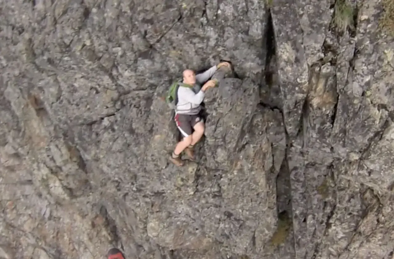 A man is climbing a steep, rocky cliff face. He's wearing shorts and a backpack, looking like he's truly "cliffed out" and facing a challenging ascent.