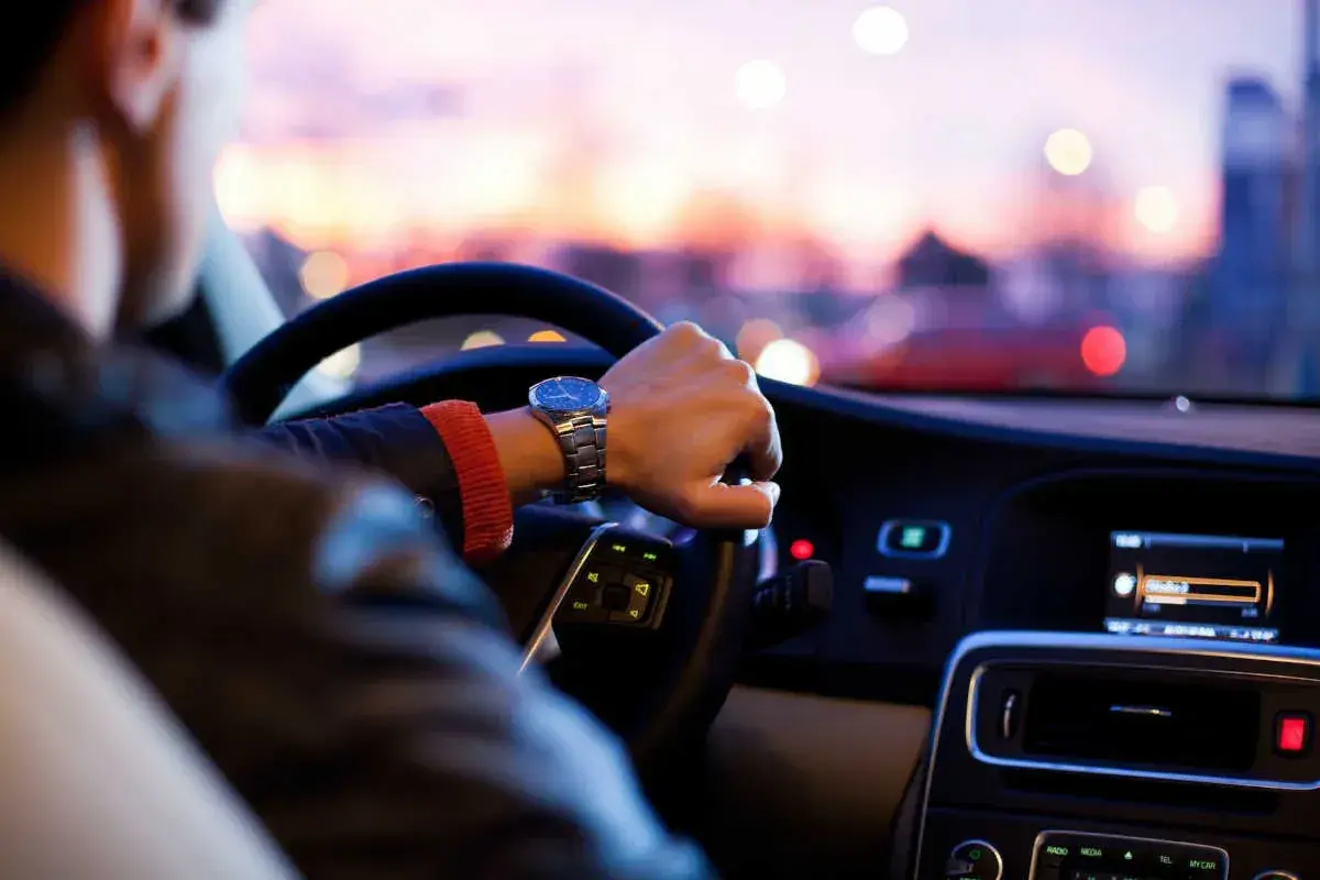 Mano al volante, orologio elegante, tramonto sfocato. Una guida esperta affronta la sera con sicurezza.
