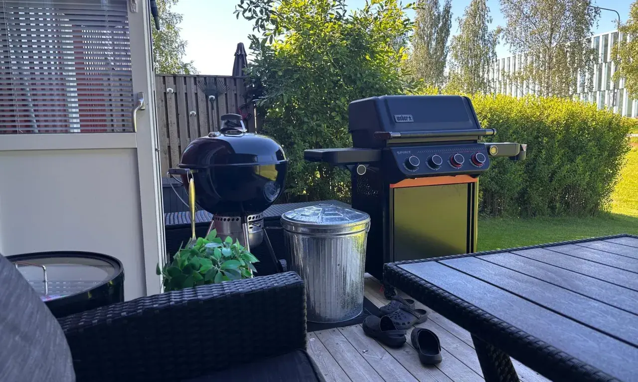 En Weber kolgrill bäst i test står redo för sommarens grillkvällar bredvid en gasolgrill. Trädgårdsmöbler och en fin gräsmatta skapar en härlig atmosfär.