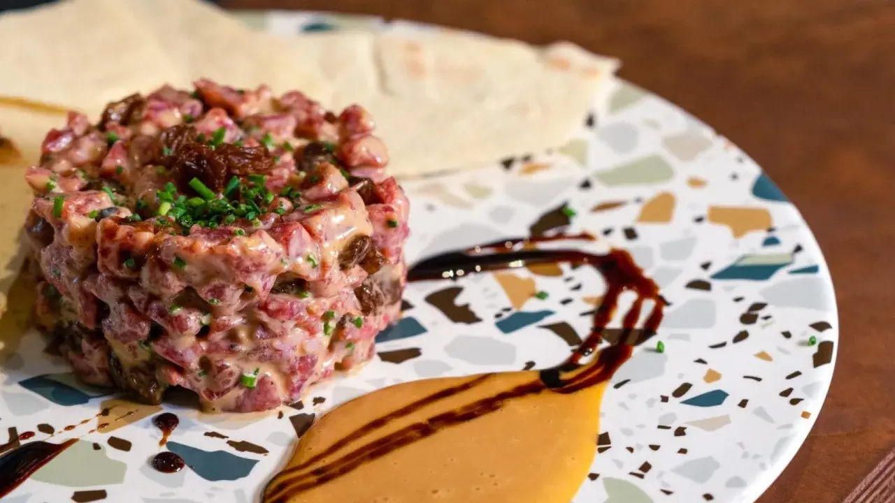 Delicioso tartar de salchichón, con trocitos de frutos secos y cebollino fresco, servido en un plato moderno con salsas.