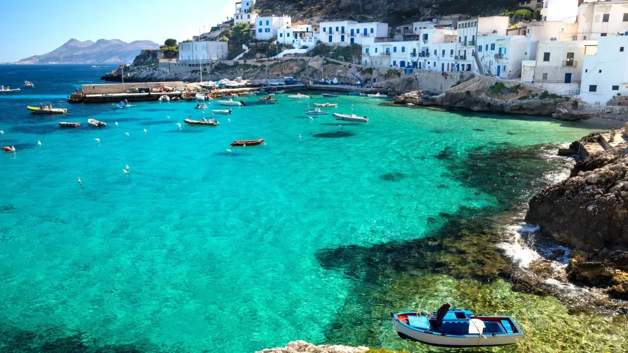 Boats in a turquoise bay near white buildings, a picturesque scene reminiscent of the charm found near international airports in Sicily.