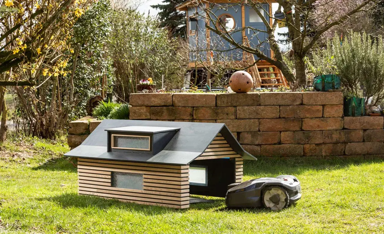 Ein moderner, selbstgebauter Rasenroboter-Unterstand mit Holzlamellen und Dachfenstern. Der Rasenroboter parkt daneben.