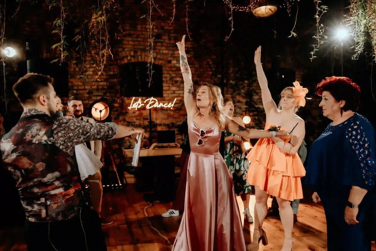 Oczepiny co to? Zabawa weselna w pełni! Panie w sukniach i panowie bawią się przy neonowym napisie "Let's Dance!".