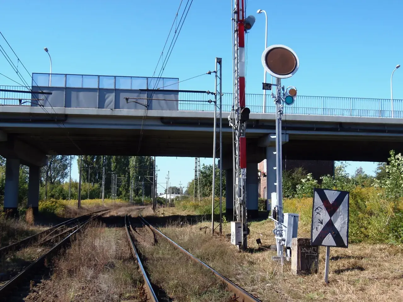Tarcza ostrzegawcza przy torach kolejowych pod wiaduktem. Sygnalizacja świetlna i znak krzyżowy informują o zbliżającym się niebezpieczeństwie.