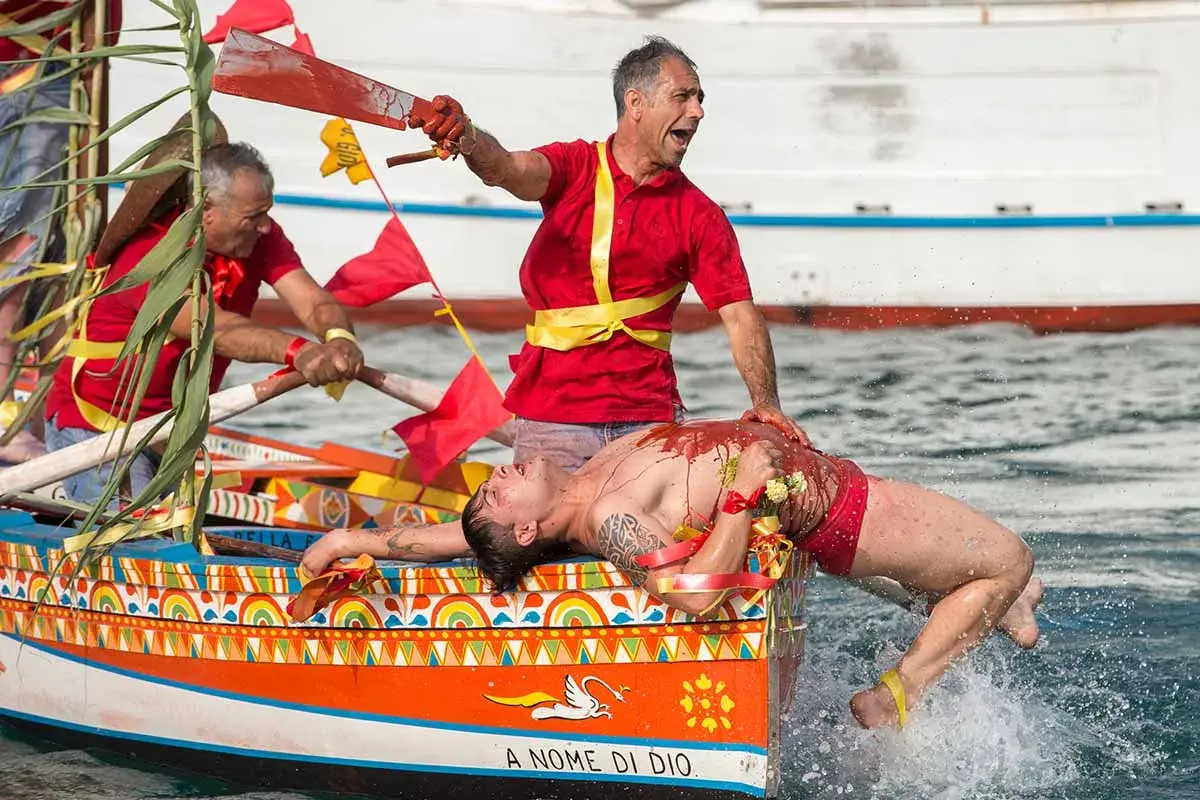 Uomo in barca siciliana, coperto di rosso, durante le feste. Un rematore con remo insanguinato.