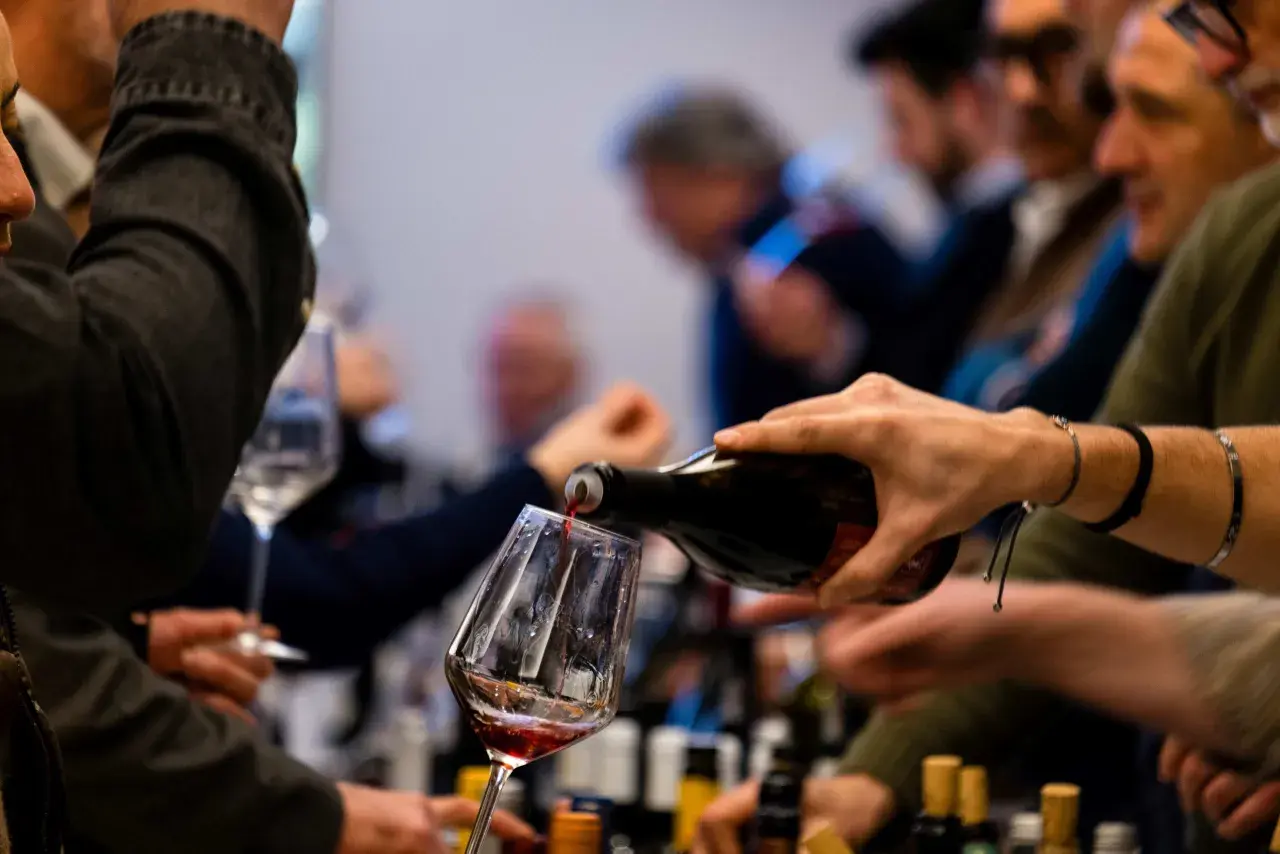 Versando vino rosso in un calice, si apprezzano le caratteristiche del vermentino. Persone in attesa, bottiglie sullo sfondo.