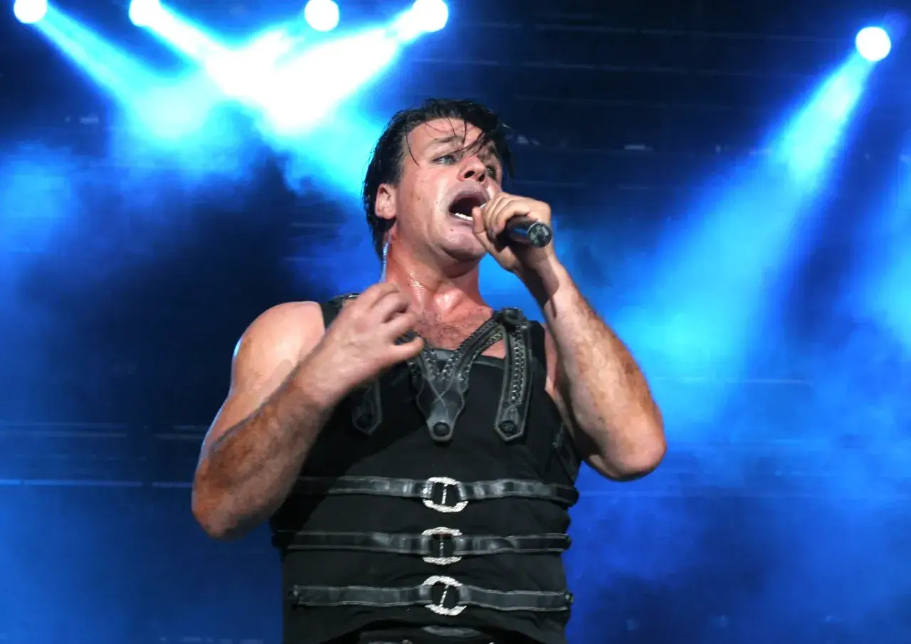 Till Lindemann na koncercie, w czarnym, skórzanym stroju, śpiewa do mikrofonu na tle niebieskich świateł.