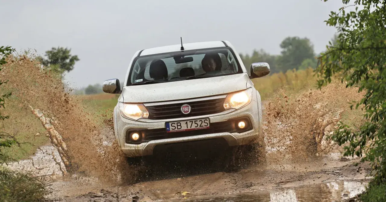 Biały Fiat Fullback z impetem wjeżdża w błotnistą kałużę, wzbijając fontanny brudu.