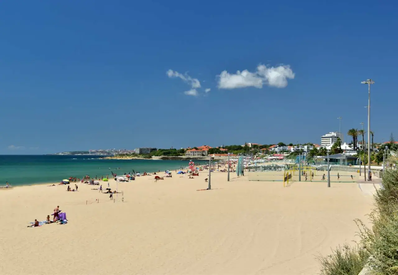 Amplia playa de arena dorada cerca de Lisboa, con gente disfrutando del sol y el mar. Hay una cancha de voleibol y edificios al fondo.