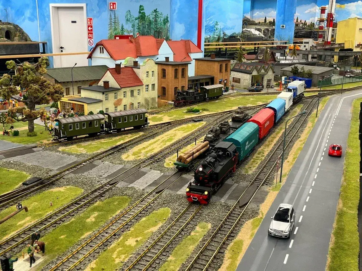 Miniaturowa kolejka z parowozem i wagonami, jak w Energylandii, przemierza malowniczy krajobraz z domkami i drogą.