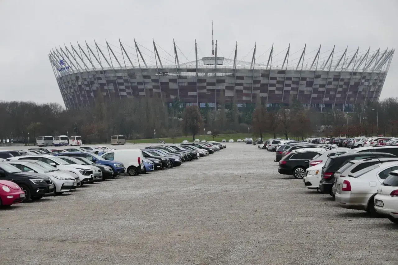 Duży parking przed Stadionem Narodowym w Warszawie. Widać rzędy samochodów i autobusów na błoniach stadionu.