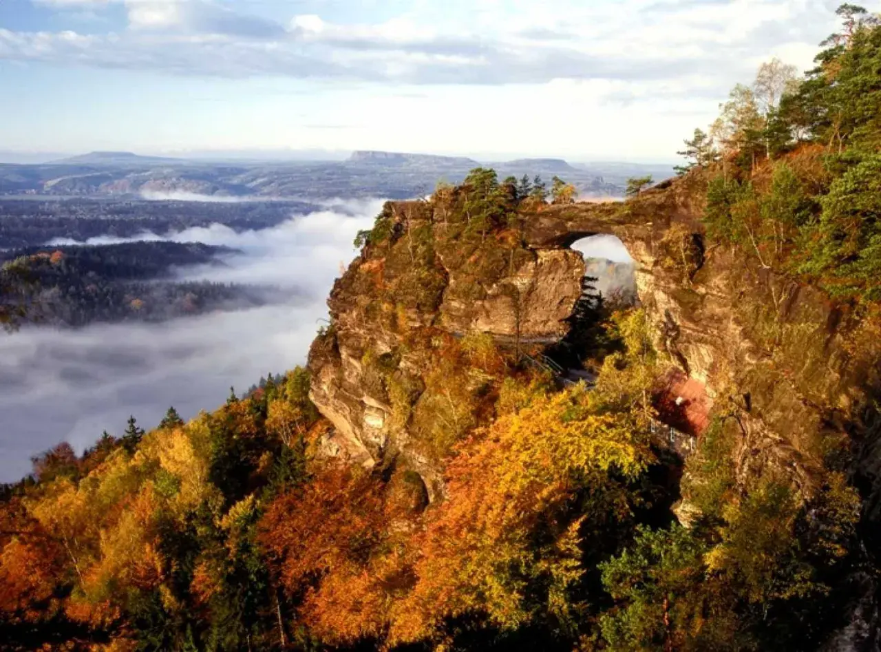 Majestätische Felsformation Pravcicka brana mit einem natürlichen Torbogen, umgeben von herbstlichem Laub und Nebel im Tal.