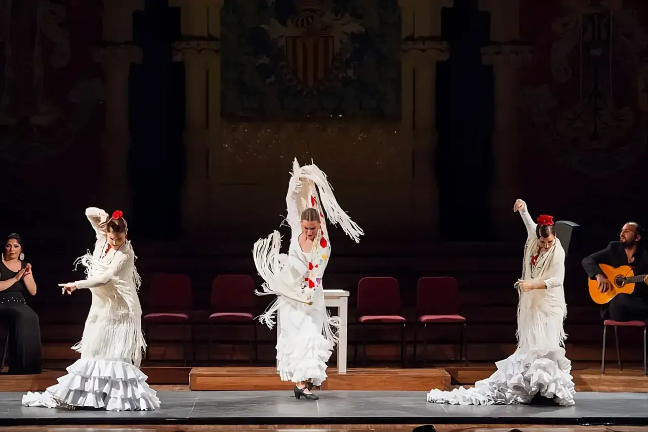 Mujeres en un apasionado baile flamenco, con trajes blancos y volantes, acompañadas por un guitarrista.