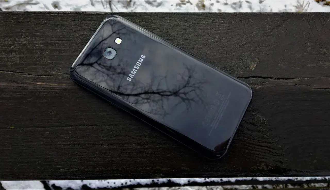 Czarny Samsung SM-A520F leży na drewnianej ławce, odbijając gałęzie drzew.