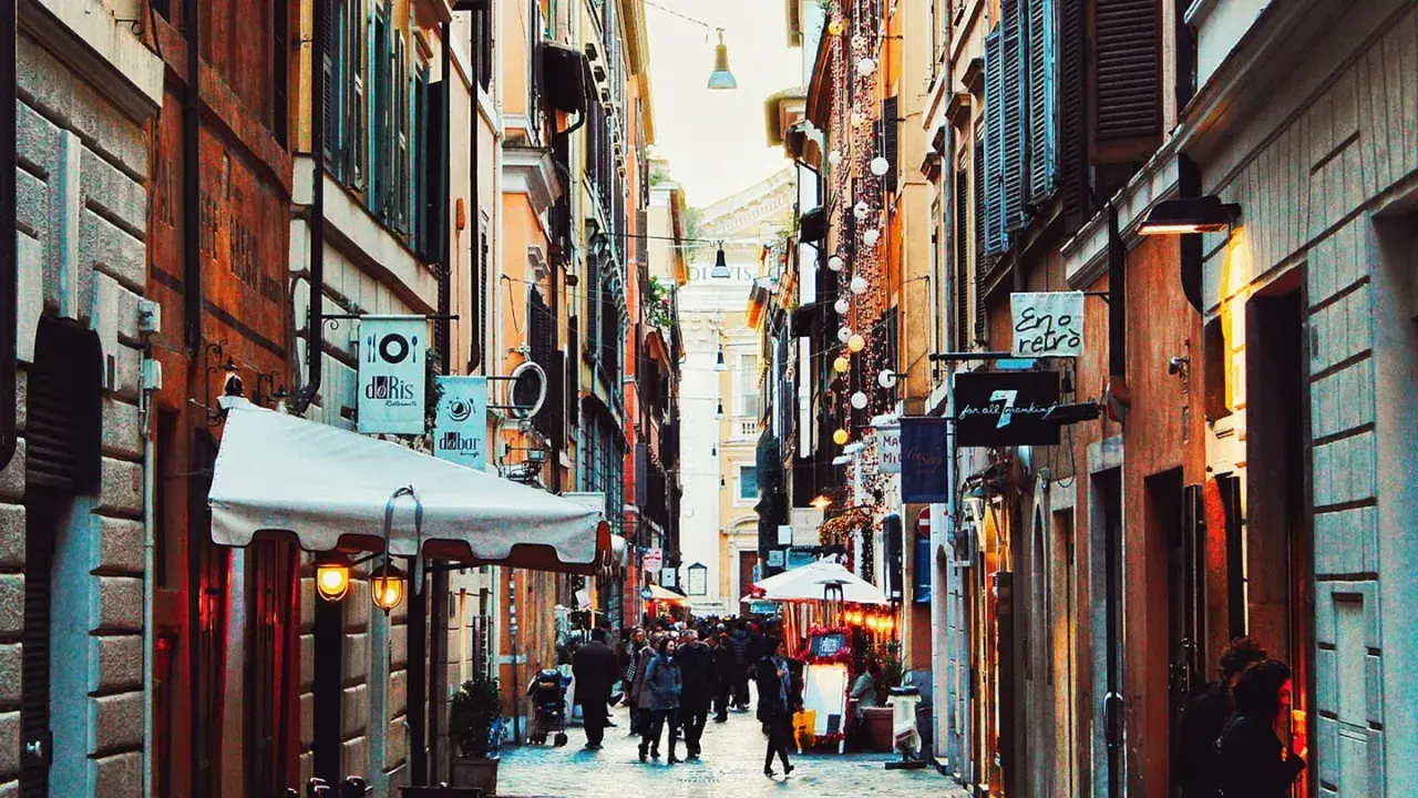 En livlig gränd i ett kvarter i Rom, med butiker och restauranger längs sidorna. Folk promenerar och njuter av atmosfären.
