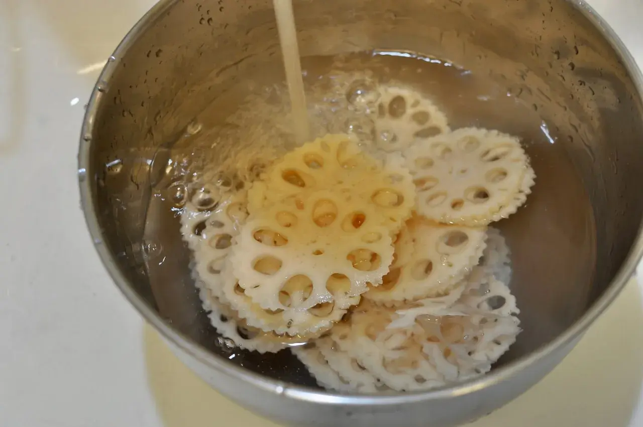Slices of pickled lotus root are being rinsed in a metal bowl under running water, ready for a delicious meal.