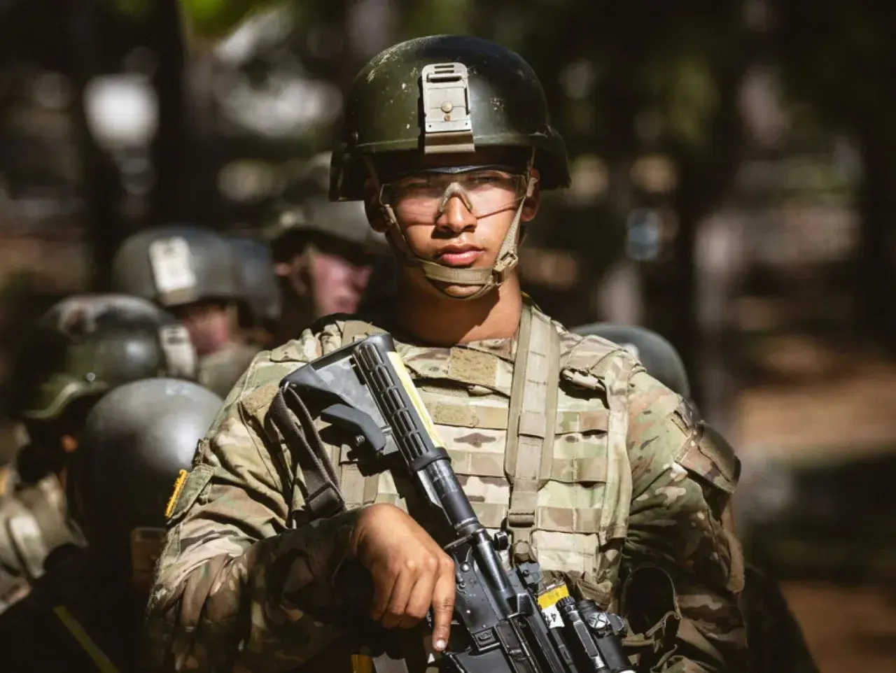 A young soldier, determined and focused, stands ready during far bootcamp. He wears a helmet, protective eyewear, and camouflage fatigues, holding his rifle.