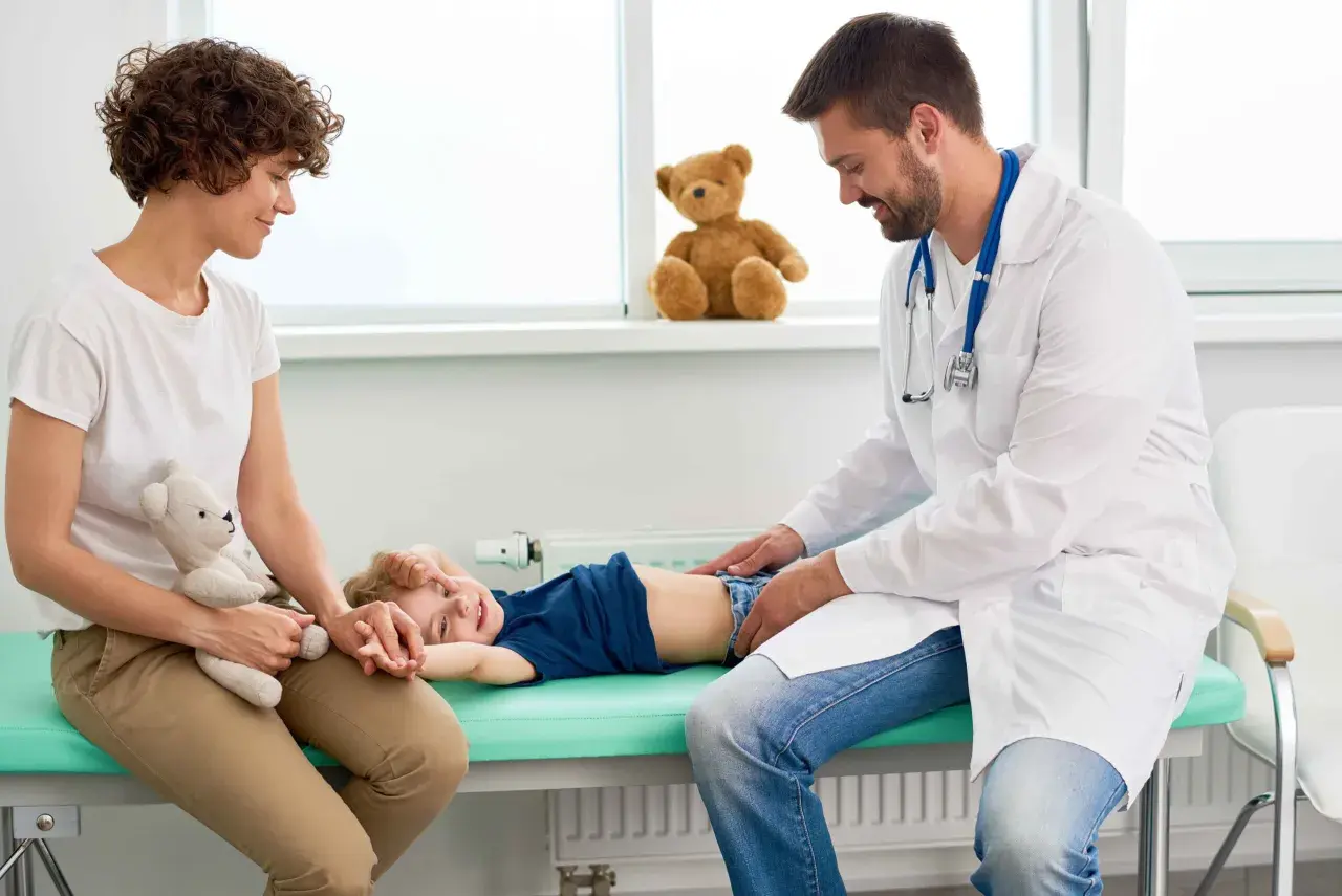 Lekarz bada brzuszek dziecka, które leży na kozetce. Mama z pluszowym misiem obok. Dziecko uśmiecha się, mimo że może mieć biegunkę u niemowlaka.