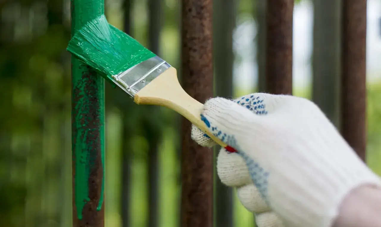 Mano guantata stende vernice verde su un palo di ferro arrugginito. La temperatura ideale per verniciare il ferro è fondamentale per una buona resa.