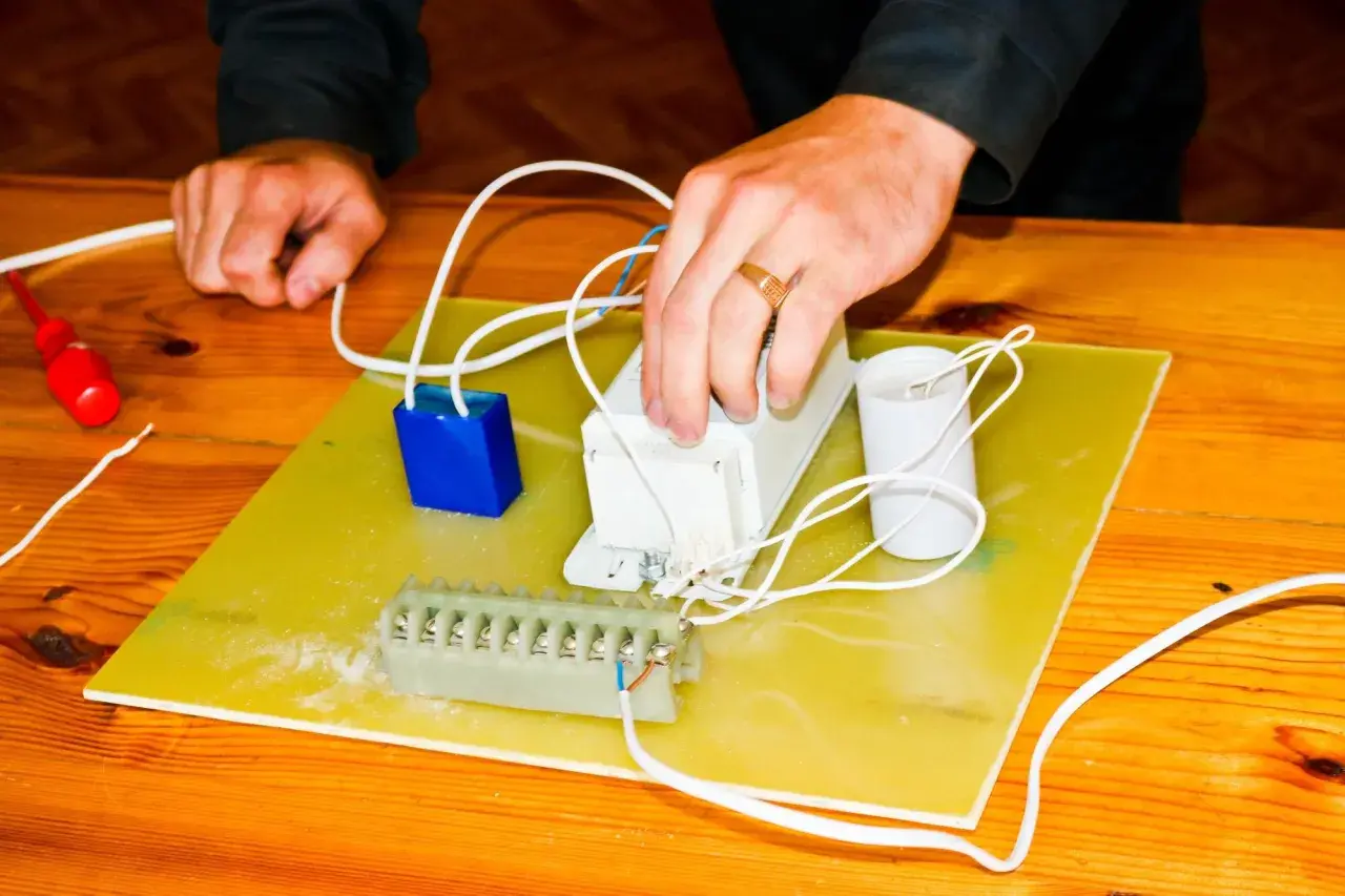 Ręce montują elementy elektryczne, przygotowując się do tego, jak podłączyć pastucha. Widać kondensator, dławik i listwę zaciskową.