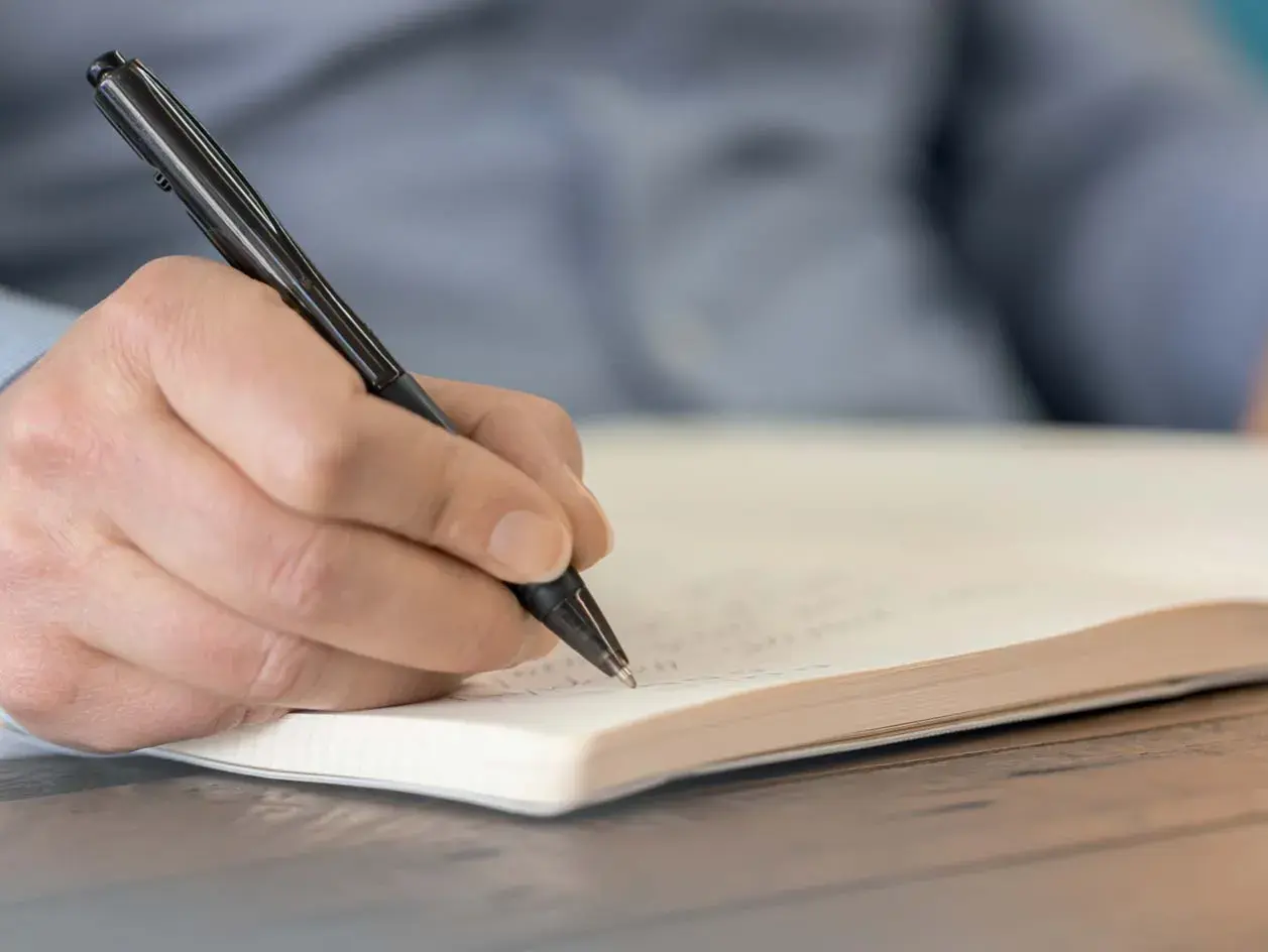 Hand schreibt mit einem Stift in ein Notizbuch. Wichtige Details für das protokoll schreiben festhalten.