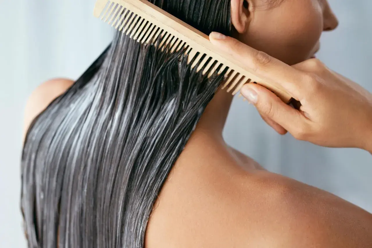 Persona peina su cabello oscuro con mascarilla y un peine de madera. Averigua cuanto tiempo dejar la mascarilla en el pelo para un tratamiento óptimo.