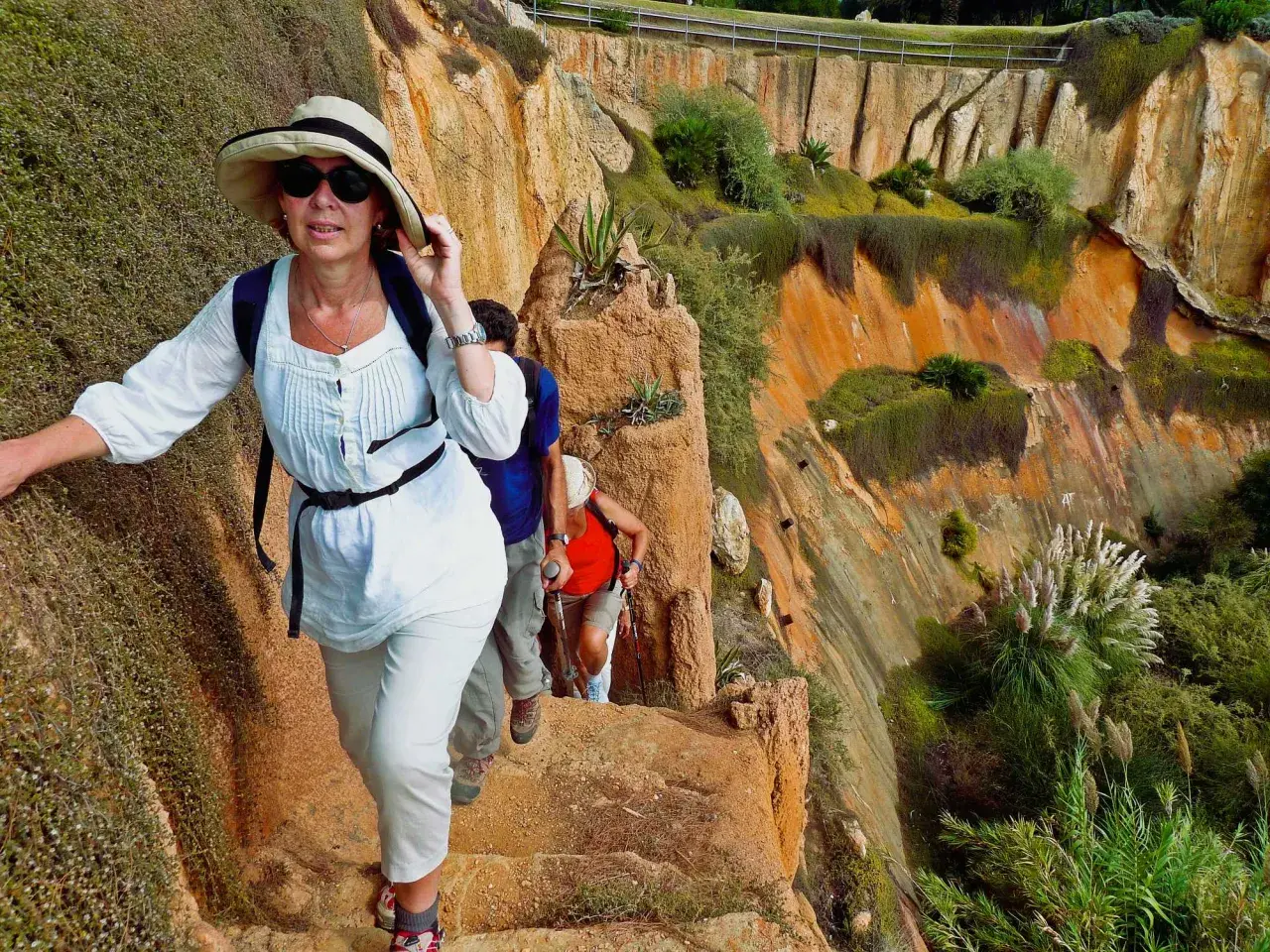 Hikers ascend a rocky path during hiking trips in Portugal, with lush green vegetation clinging to the orange cliffs.