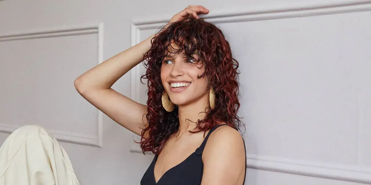 Donna sorridente con capelli castani mossi e tinta rossa, indossa orecchini dorati.