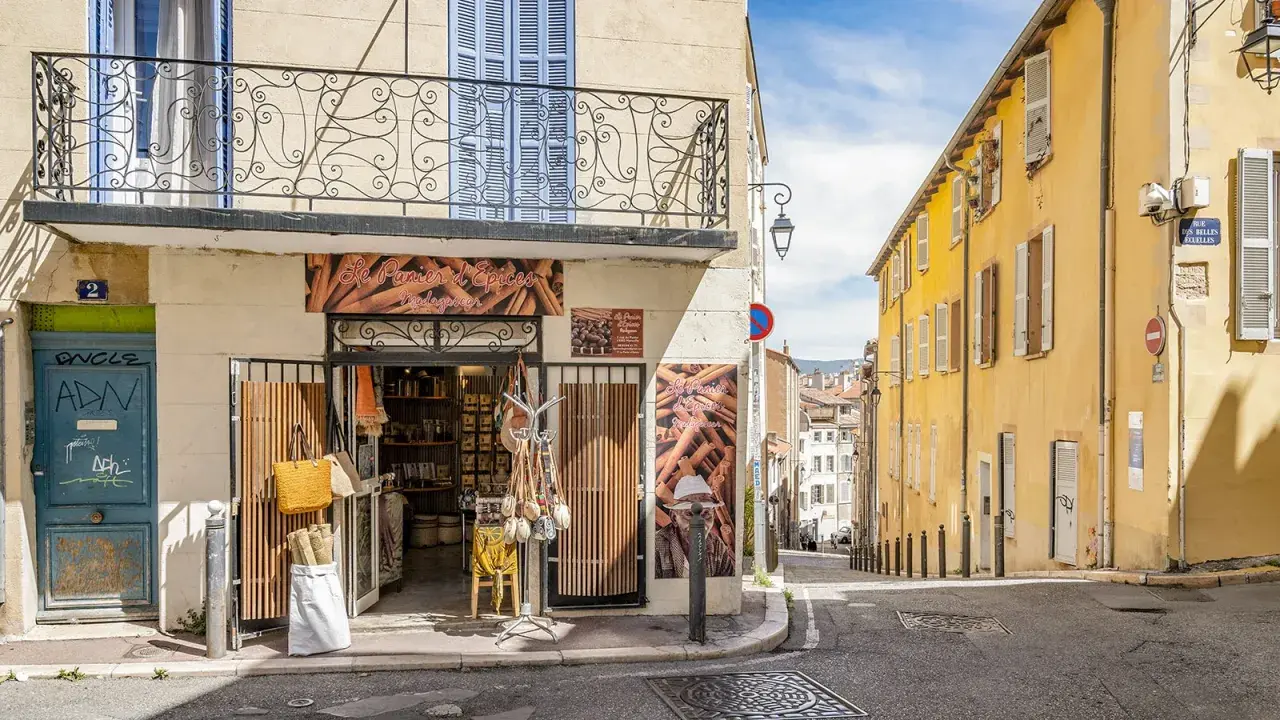 Bunte Häuser und ein Gewürzladen in der **Marseille Altstadt**. Eine schmale Gasse führt bergauf, gesäumt von sonnengelbem Mauerwerk und blauen Fensterläden.