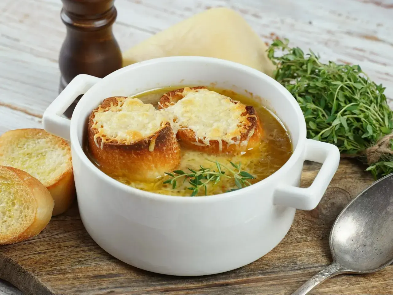 Aksamitna zupa cebulowa przepis, podana w białej miseczce, z grzankami zapieczonymi z serem i gałązką tymianku. Obok kromki chleba.