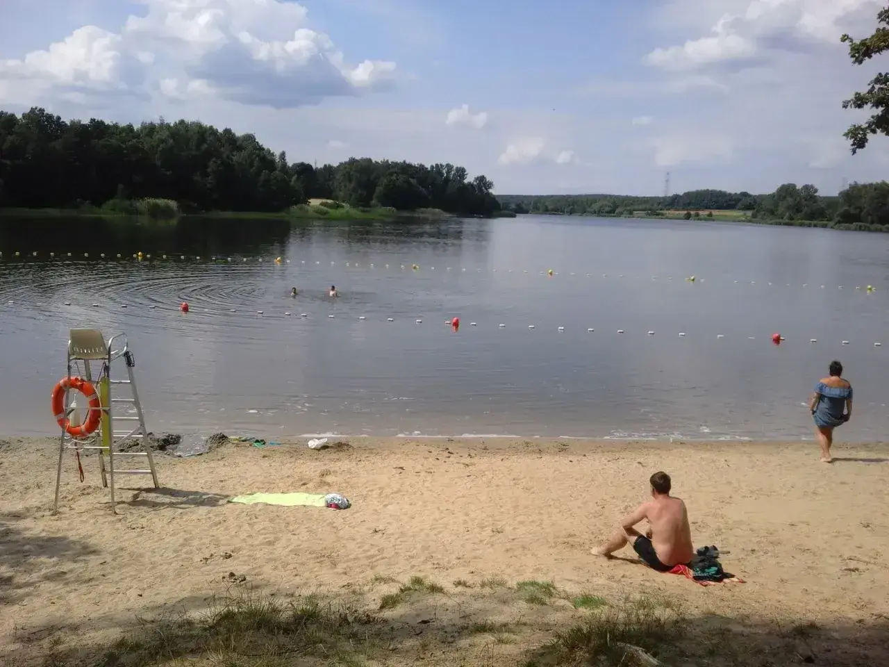 Słoneczny dzień nad rybnikiem zalew. Kąpielisko z bojami, ratownik na posterunku, plażowicze odpoczywają na piasku.
