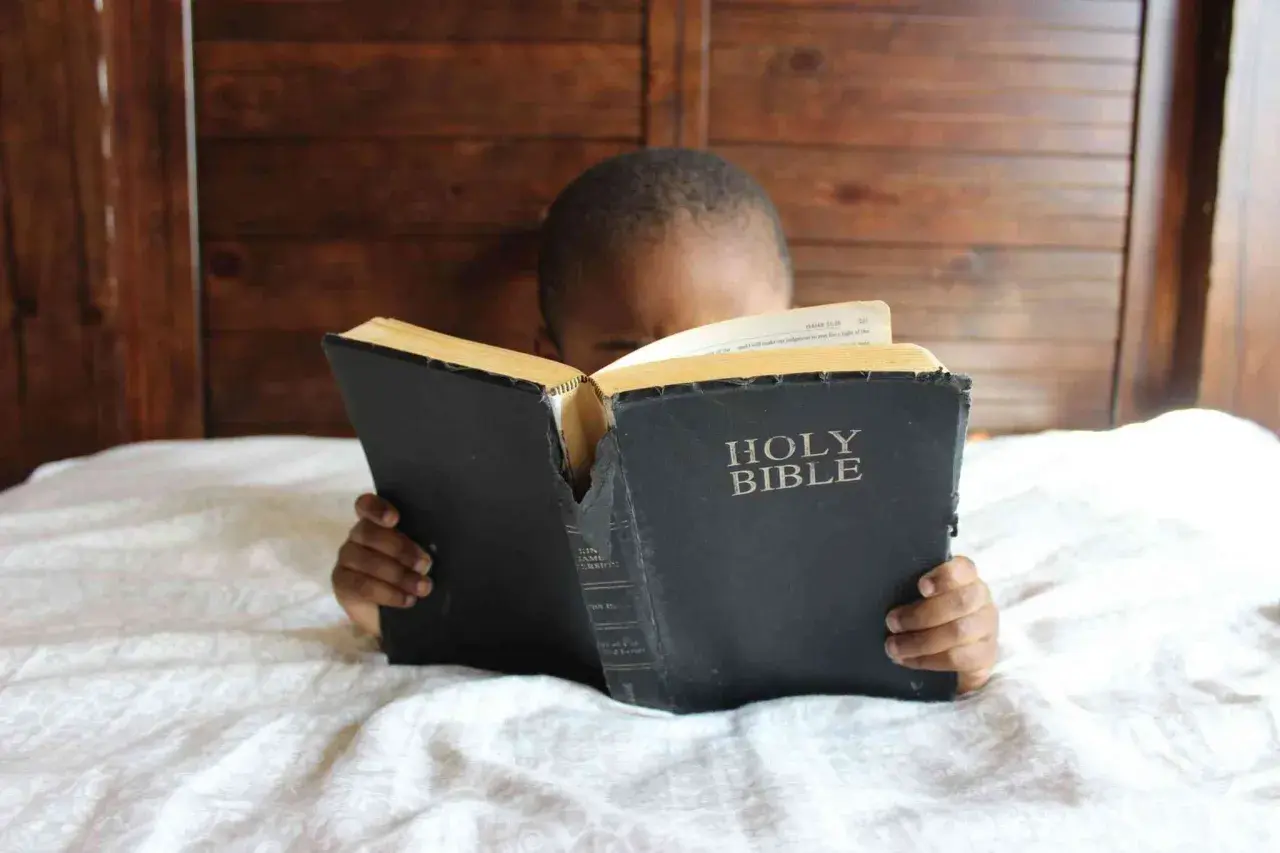 Dziecko z zaciekawieniem czyta Biblię, leżąc w łóżku. Słowa "Holy Bible" są widoczne na okładce.