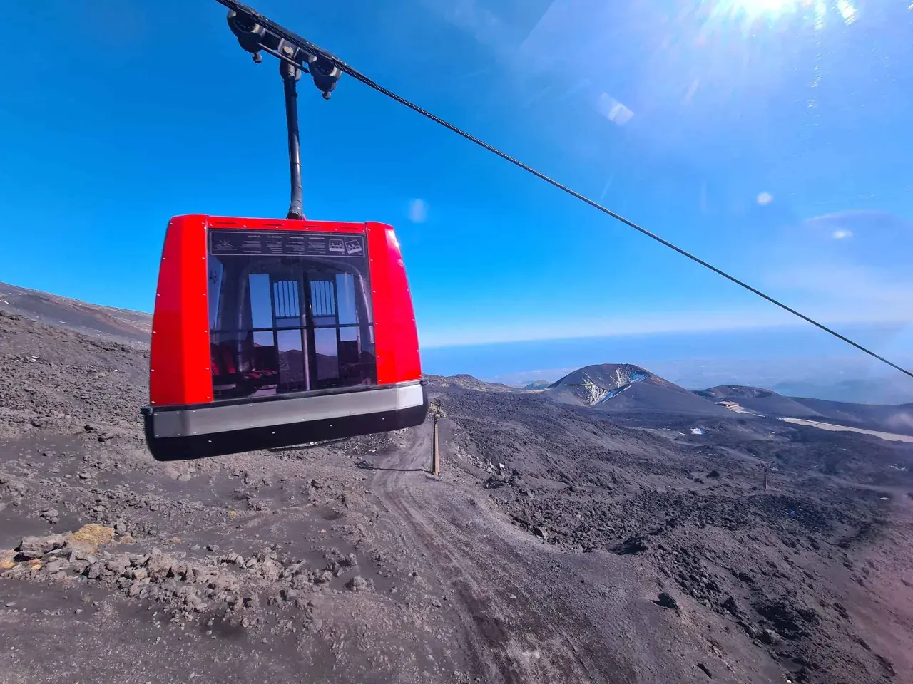 Cabina rossa della funivia in viaggio verso l'altezza Etna Rifugio Sapienza, su un paesaggio lunare di rocce laviche.