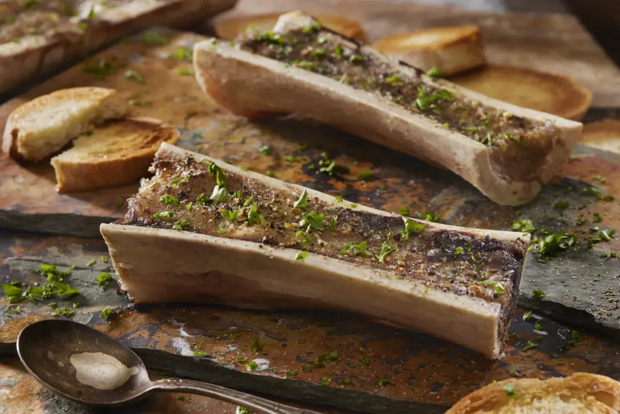 Deux os à moelle rôtis, parsemés de persil et de poivre, accompagnés de toasts grillés. Une cuillère attend patiemment.