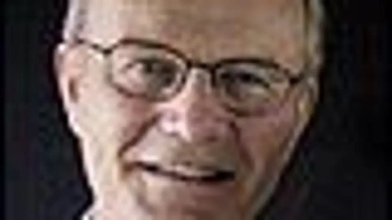 Närbild på Olle Stenholm med glasögon, ett vänligt leende och mörkt bakgrund.