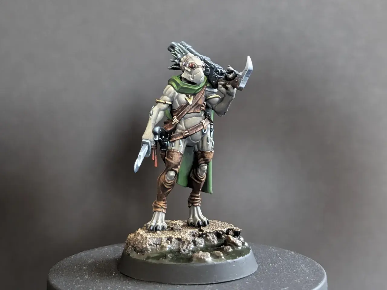 Zbrojny kroot w zielonej pelerynie i brązowych ochraniaczach, z nożem w dłoni i karabinem na plecach, gotowy do walki.