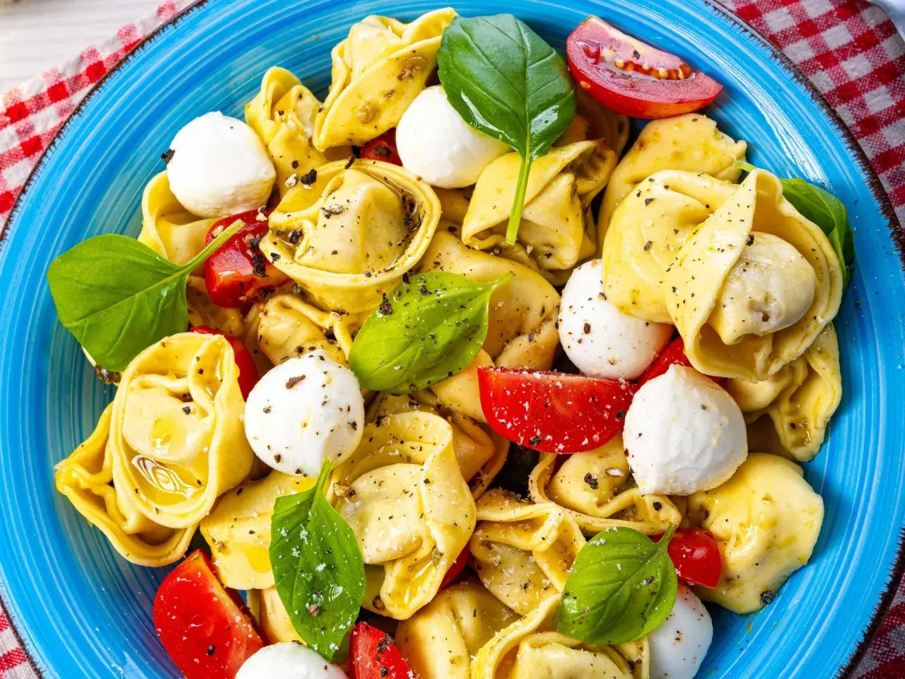 Ein bunter Tortellini Salat mit Mozzarella-Kugeln, Tomatenstücken und frischem Basilikum, appetitlich in einer blauen Schüssel angerichtet.